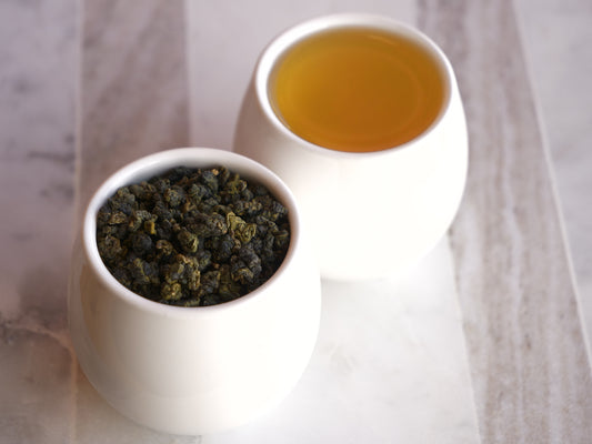 Two white ceramic cups, one filled with green tea leaves and the other with brewed tea, on a light wooden surface.