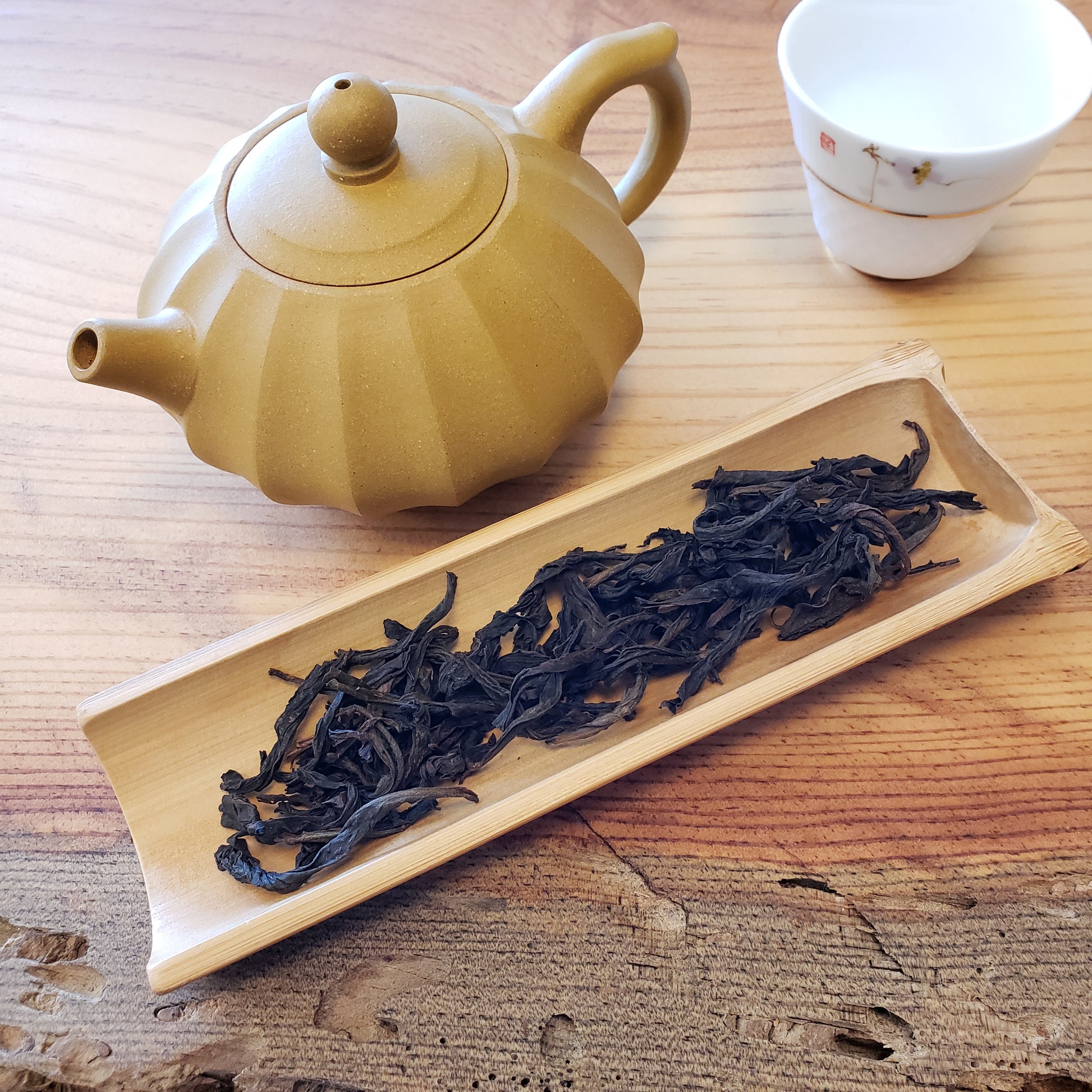 Yellow teapot next to a wooden tray with dried tea leaves on a wooden surface.