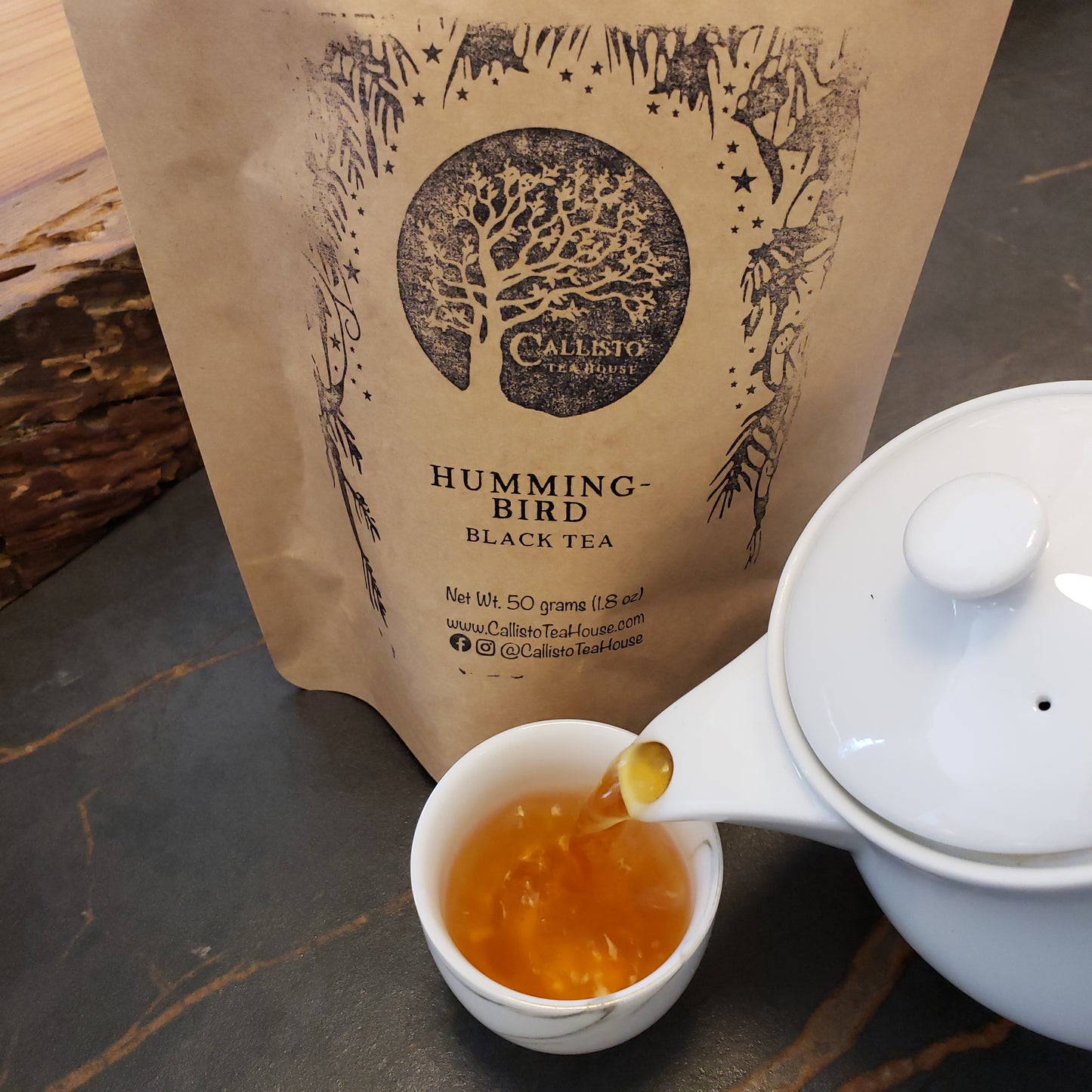 White teapot pouring tea into a small white cup next to a 'Hummingbird Black Tea' package.