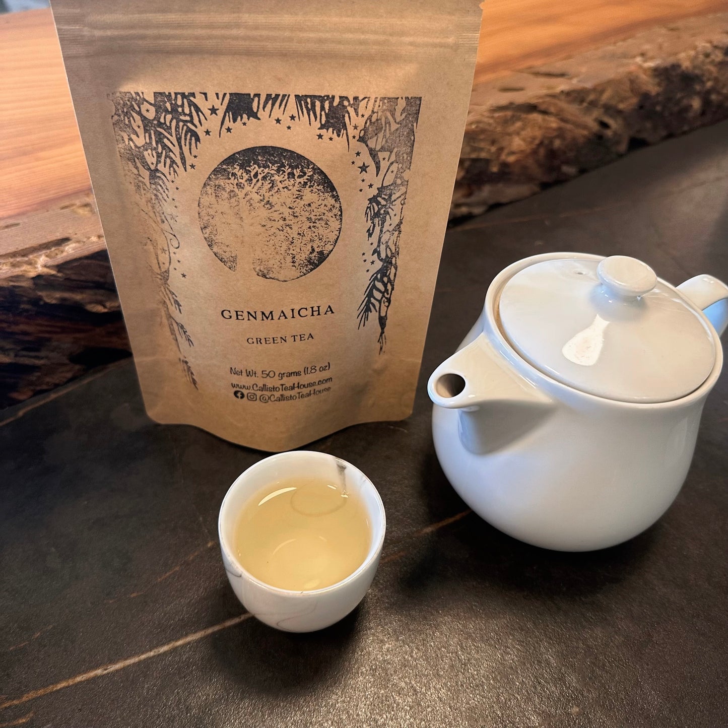 White teapot with a white cup filled with tea in front of a package of Genmaicha tea