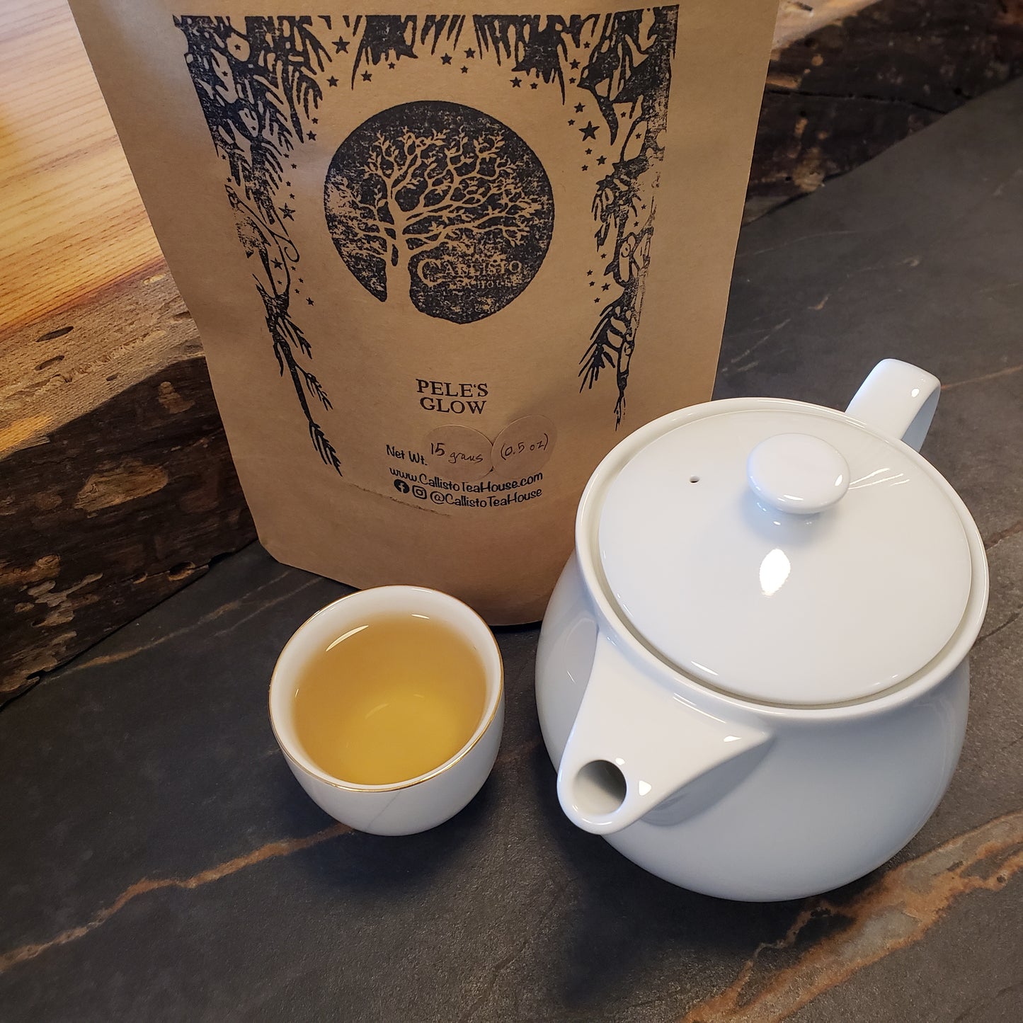 White teapot and cup with brewed tea on a wooden surface, next to a branded tea bag.