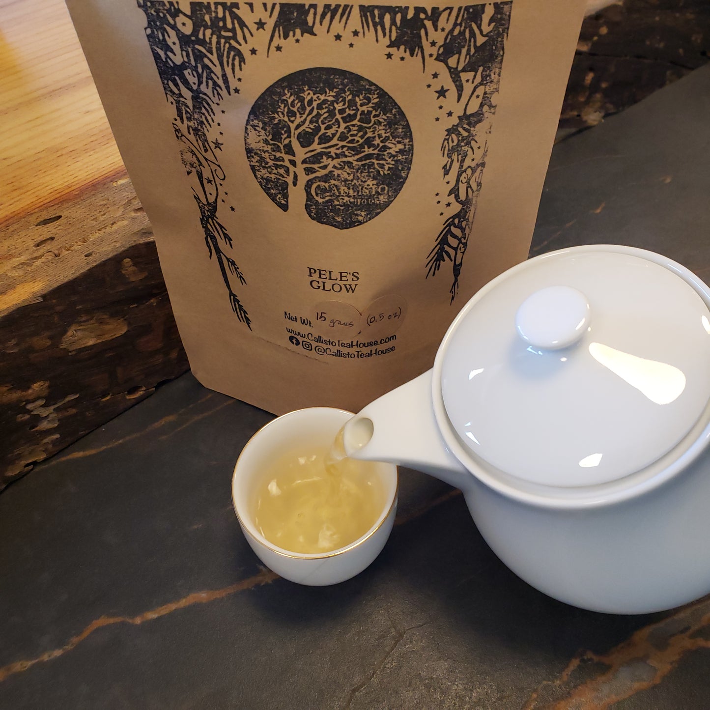 White teapot pouring tea into a white cup with a Peles Glow tea bag in the background.