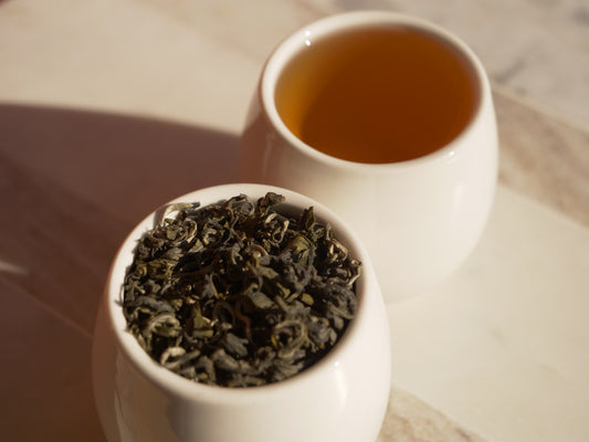 White cup with green tea leaves and a white cup with brewed green tea on a wooden surface