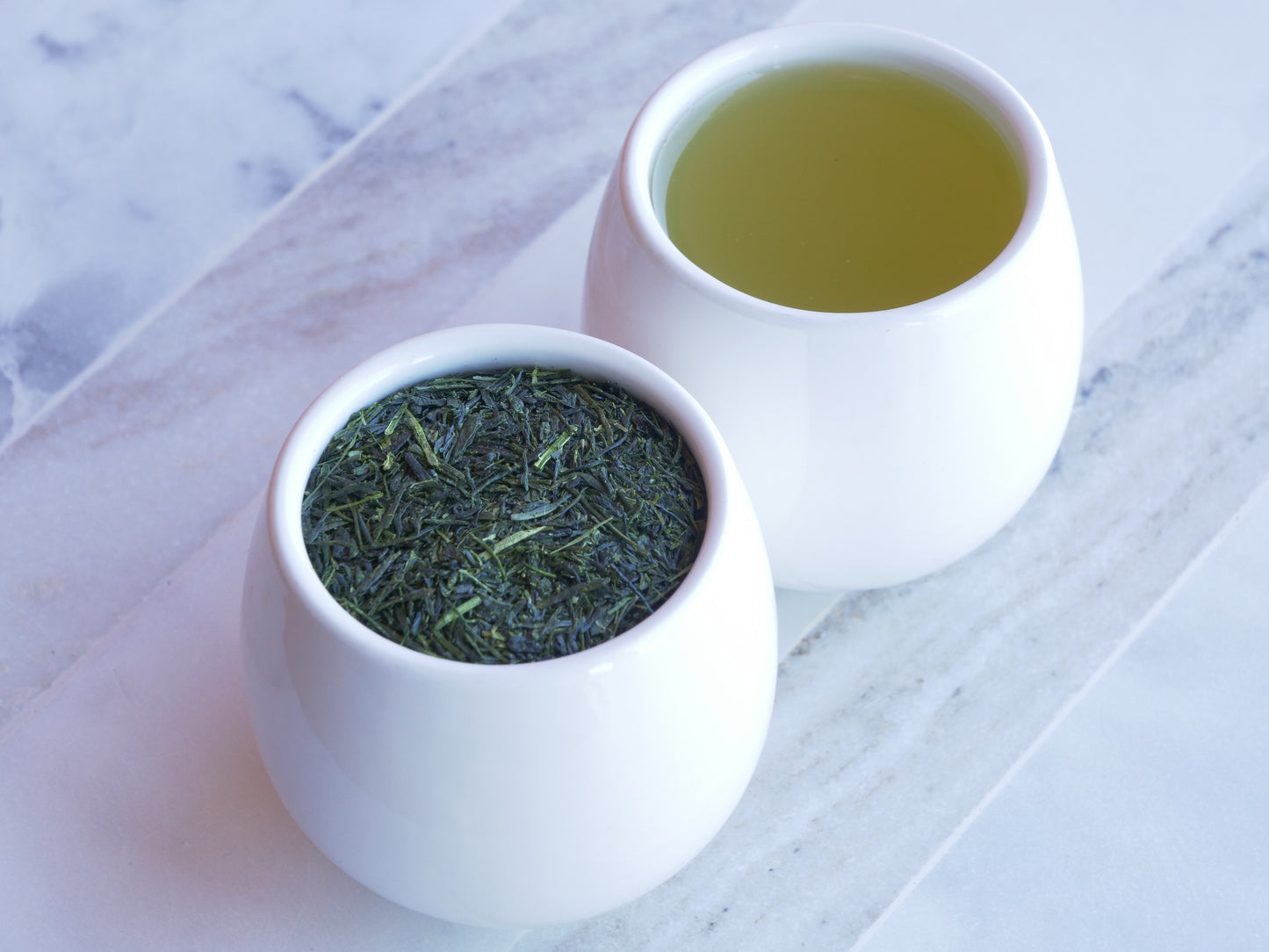 Two white ceramic bowls, one filled with green tea leaves and the other with brewed green tea, on a marble surface.