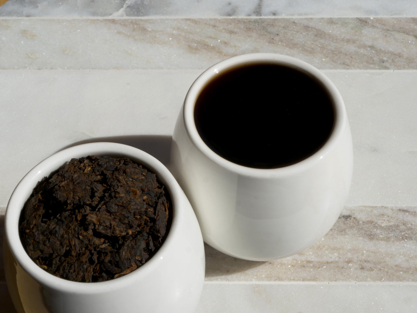 Two white cups, one filled with tea leaves and the other with brewed tea, on a marble surface.