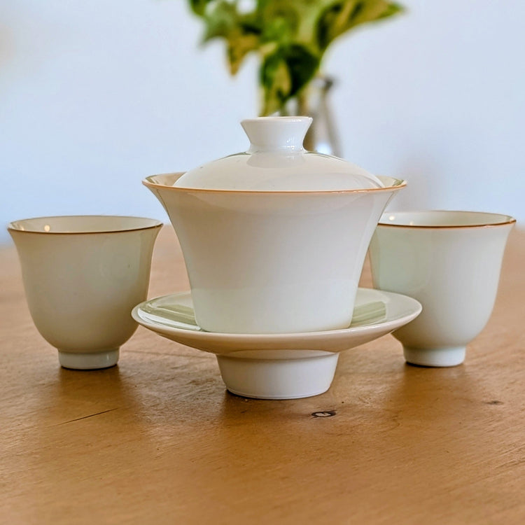 A white ceramic tea set with a lidded gaiwan on a matching saucer, flanked by two small cups, displayed on a wooden tabletop with a soft blurred background.