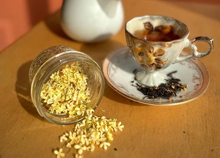A styled tea scene with a white teapot, a patterned teacup and saucer filled with tea, and a small glass jar tipped over with loose dried flowers on a wooden table against a warm terracotta background.