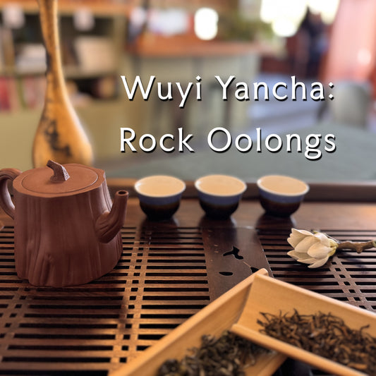 A Wuyi Yancha rock oolong tea set arranged on a dark bamboo tea tray, featuring a brown ceramic log-shaped teapot, three small tea cups, loose leaf tea in wooden trays, and a white flower, with the text 'Wuyi Yancha: Rock Oolongs' overlaid on the image.
