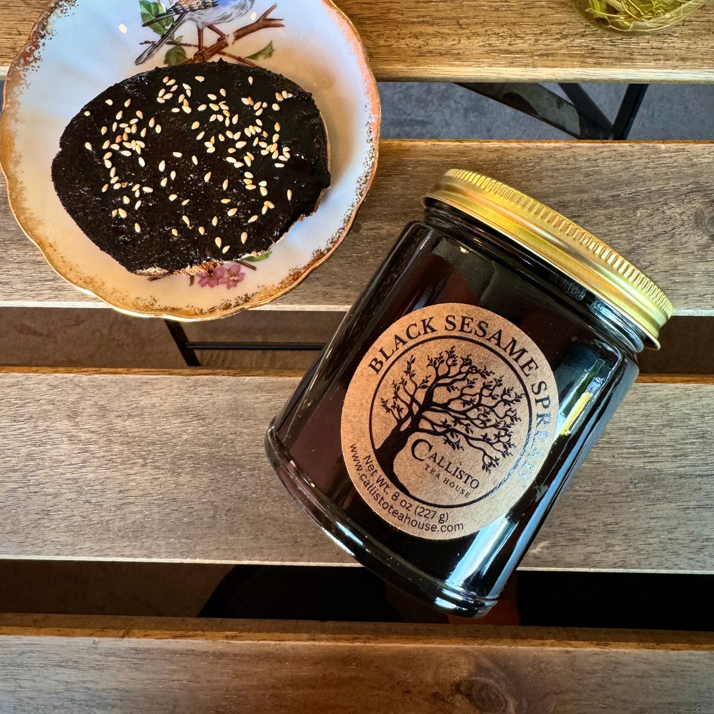 Jar of black sesame spread on a wooden surface with a decorative plate in the background.