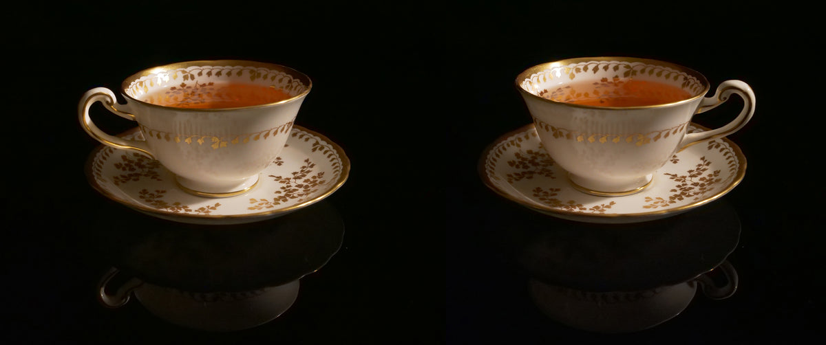 Two white porcelain teacups with gold floral detailing and gold-rimmed edges, filled with amber tea, displayed side by side on matching saucers against a black reflective background.