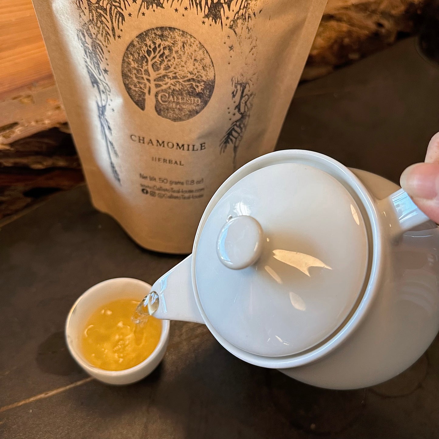 White teapot pouring tea into a small white cup with a package of chamomile tea in the background.