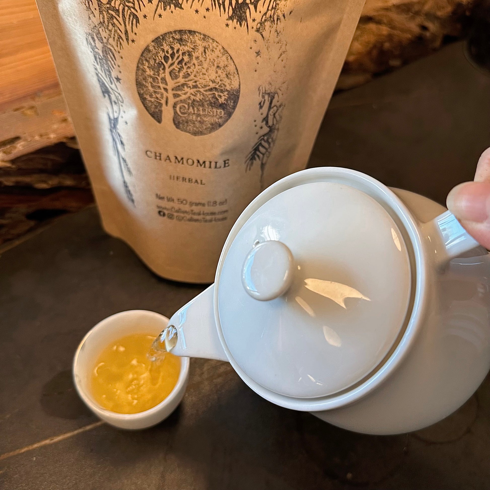 White teapot pouring tea into a small white cup with a package of chamomile tea in the background.