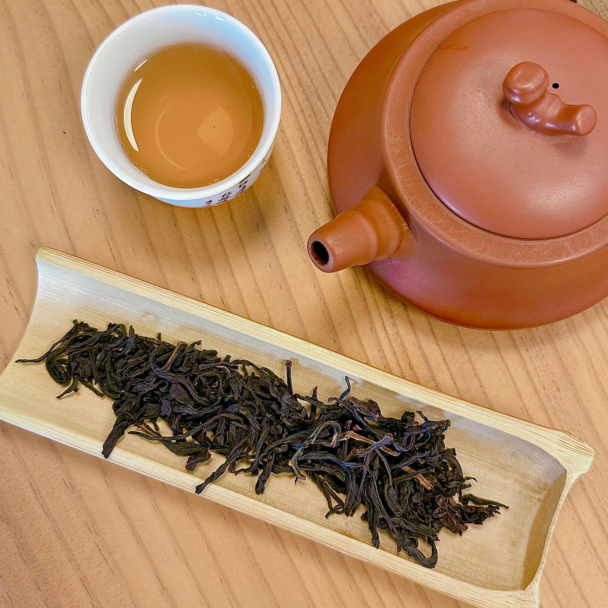 Tea set with a teapot, cup of tea, and tray of loose tea leaves on a wooden surface.