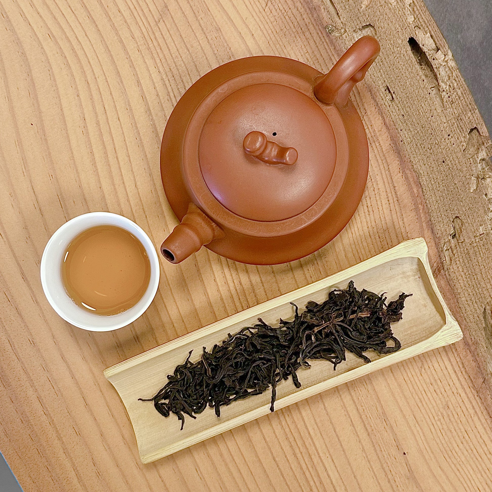 Clay teapot, white cup with tea, and wooden tray with tea leaves on a wooden surface