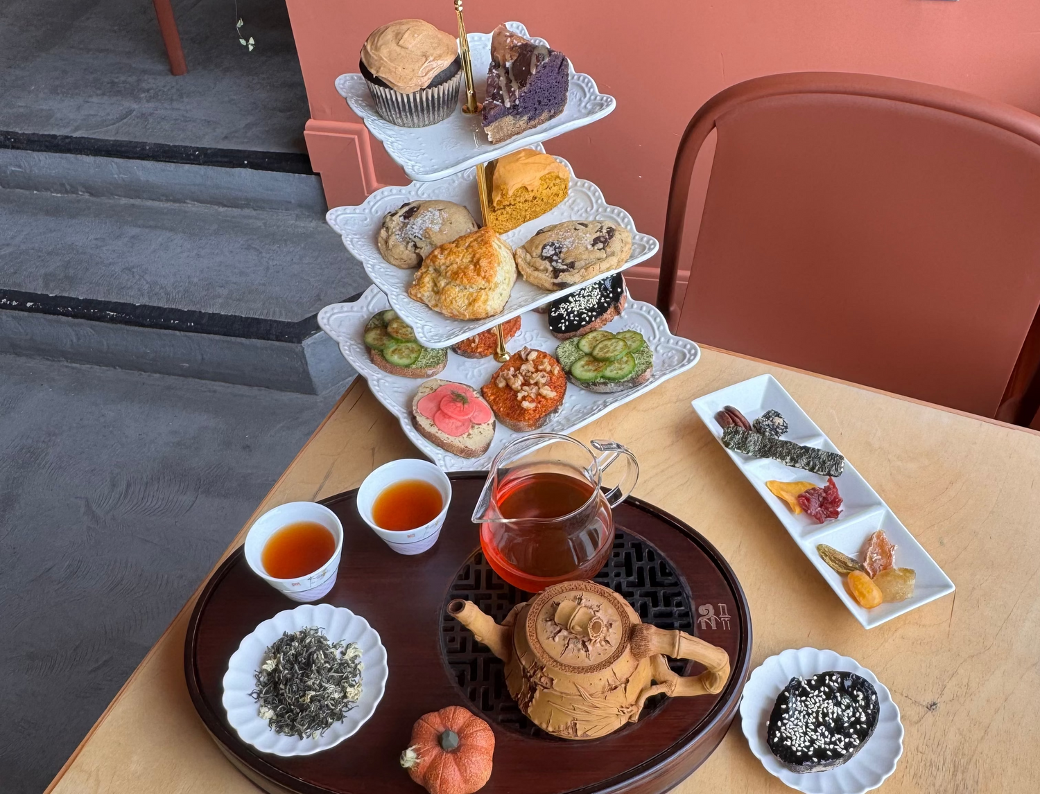 A styled tea and pastry spread on a light wood table, featuring a small wooden teapot, two cups of tea, loose-leaf tea, dried fruit, and a three-tiered stand filled with assorted cakes and pastries. The background shows a terracotta wall and a chair.