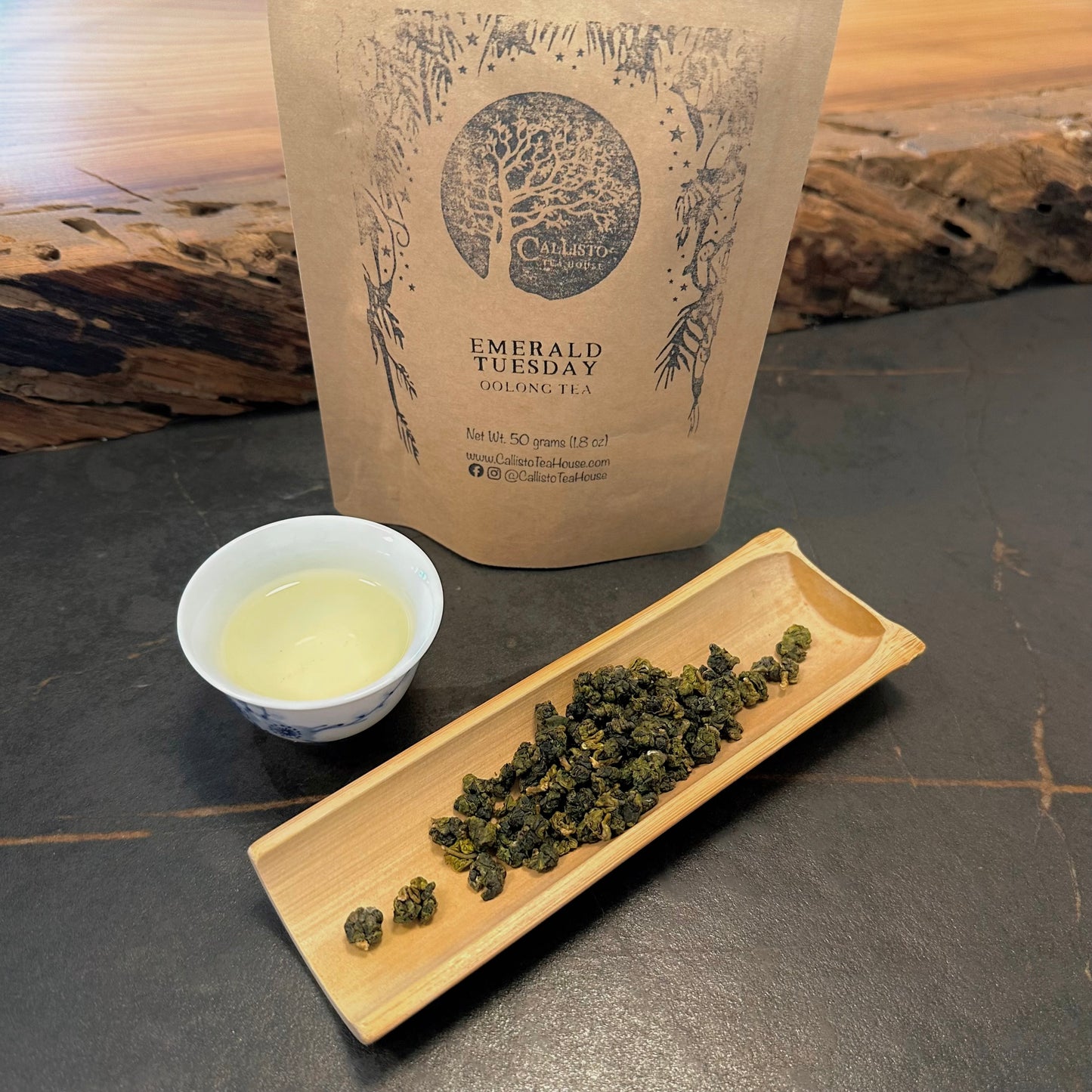 Tea packaging, tea leaves on a tray, and a small cup of tea on a dark surface.
