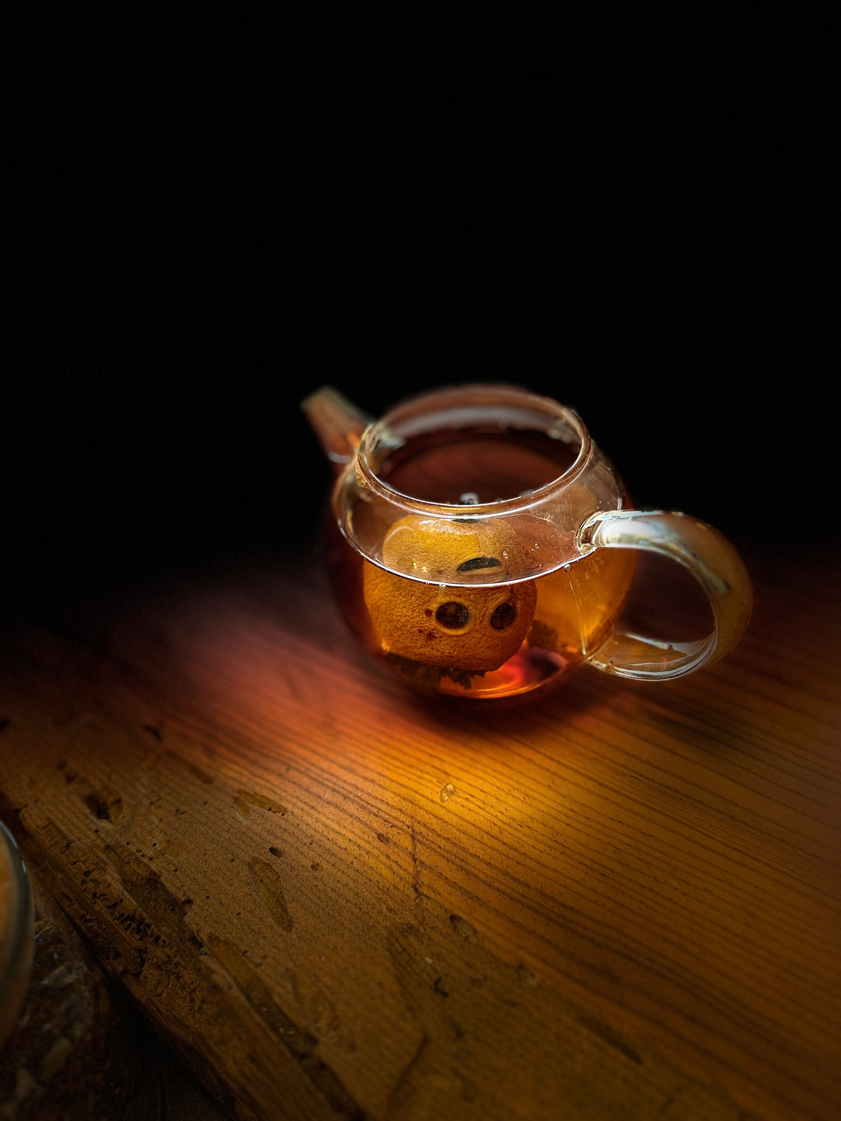 A small clear glass teapot filled with amber tea and a visible mini lemon filled with tea leaves, placed on a wooden surface against a dark background. Warm light reflects off the glass and wood.