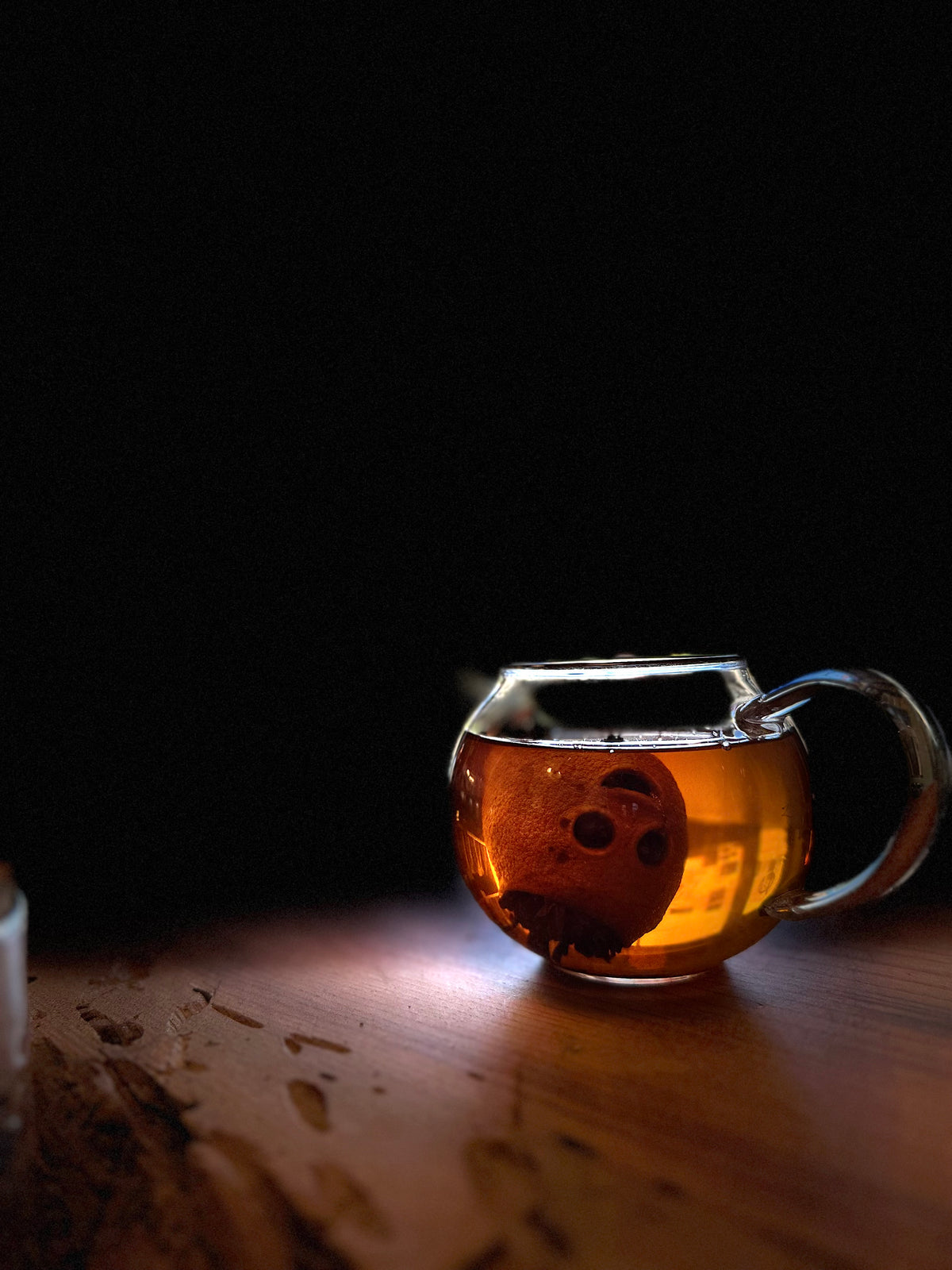 A clear glass teapot with a handle, filled with amber-brown tea and a floating tea-filled mini lemon, resting on a wooden surface against a dark background.