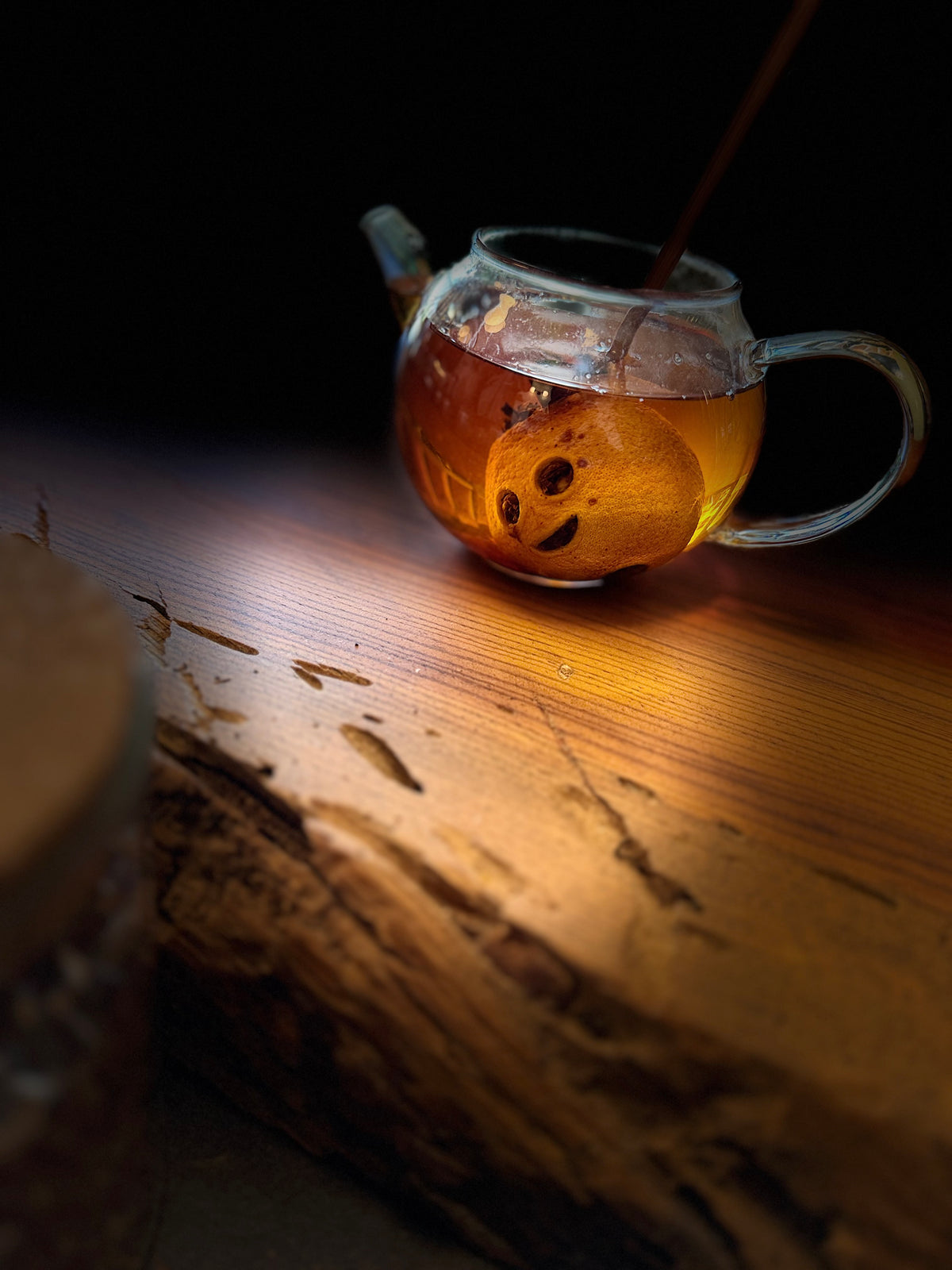 A clear glass teapot filled with amber tea and a carved mini lemon, set on a rustic wooden surface against a dark background with dramatic warm lighting.