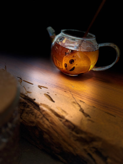 A clear glass teapot filled with amber tea and a carved mini lemon, set on a rustic wooden surface against a dark background with dramatic warm lighting.