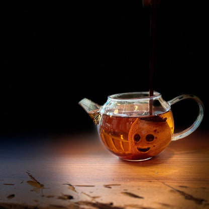 A clear glass teapot filled with amber tea and a carved mini lemon filled with tea leaves sits on a wooden surface against a dark background.