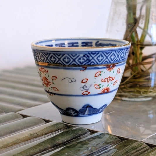 A small white ceramic cup decorated with blue geometric patterns, red floral motifs, and blue mountain designs, sitting on a reflective glass surface.