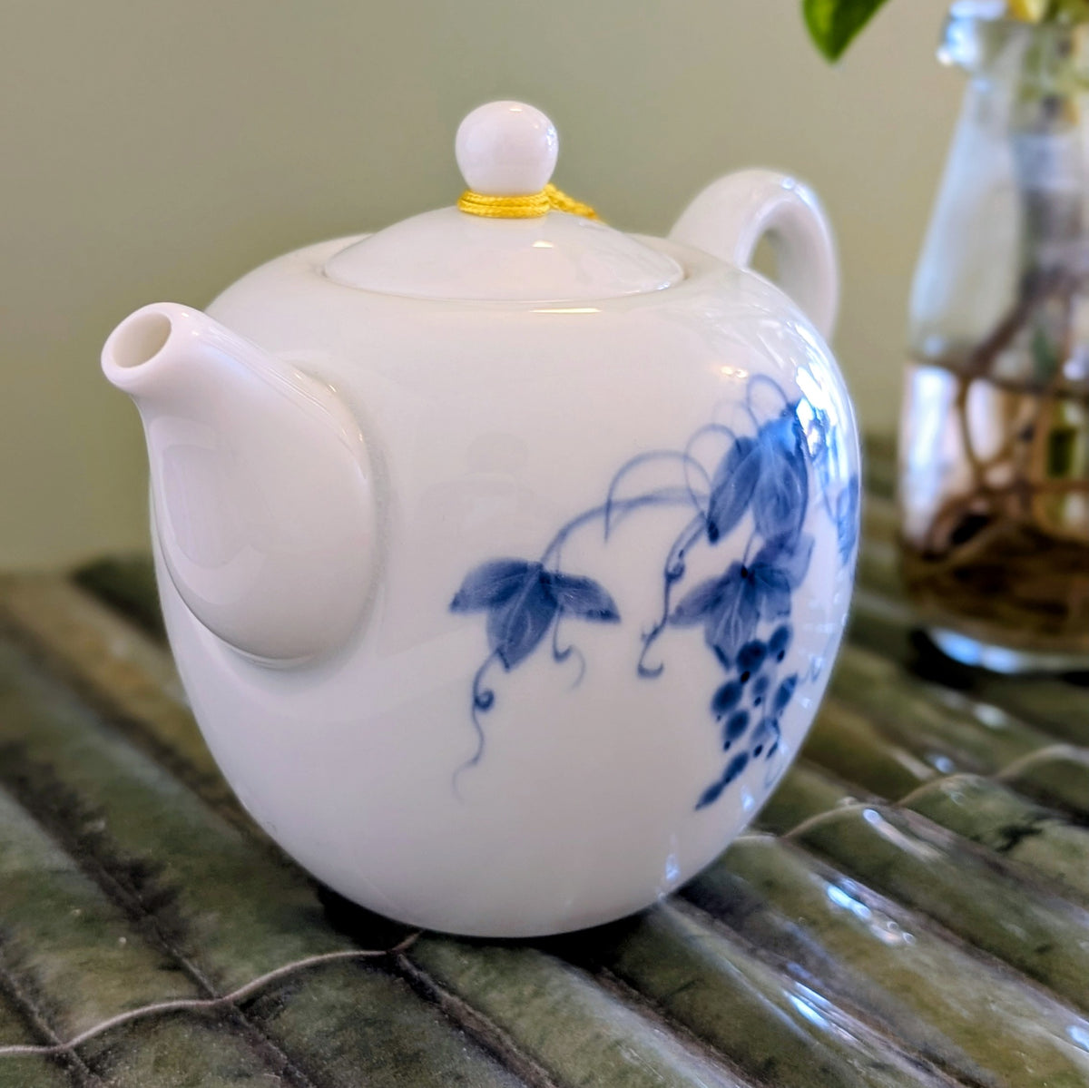 A white ceramic teapot decorated with blue floral vine patterns, resting on a green textured mat with a blurred glass vase in the background.