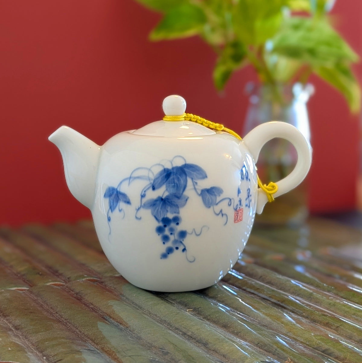 A white ceramic teapot with blue floral and vine patterns, featuring a small red seal and a yellow cord detail, resting on a textured green surface against a red background.