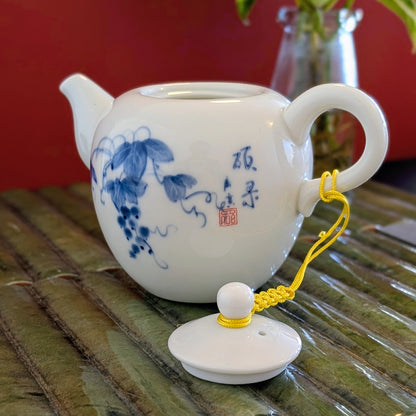 A white ceramic teapot with blue floral and calligraphic decoration, shown with its matching lid placed in front on a bamboo mat against a red background.