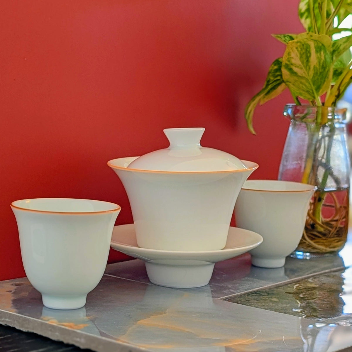 A white ceramic tea set with a lidded gaiwan and two matching cups, each trimmed with a thin ren line, arranged on a reflective gray surface against a solid red background with a glass vase of green leaves in the back.