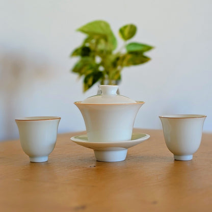 A three-piece ceramic tea set on a wooden table, featuring a central lidded teapot on a saucer with two small cups, set against a softly blurred background with a green plant.