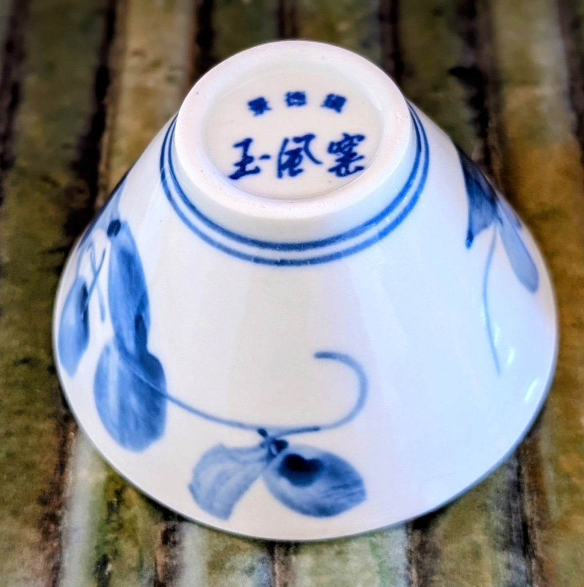 Bottom of a white porcelain tea cup with blue painted leaf design and Chinese characters on a wooden mat.