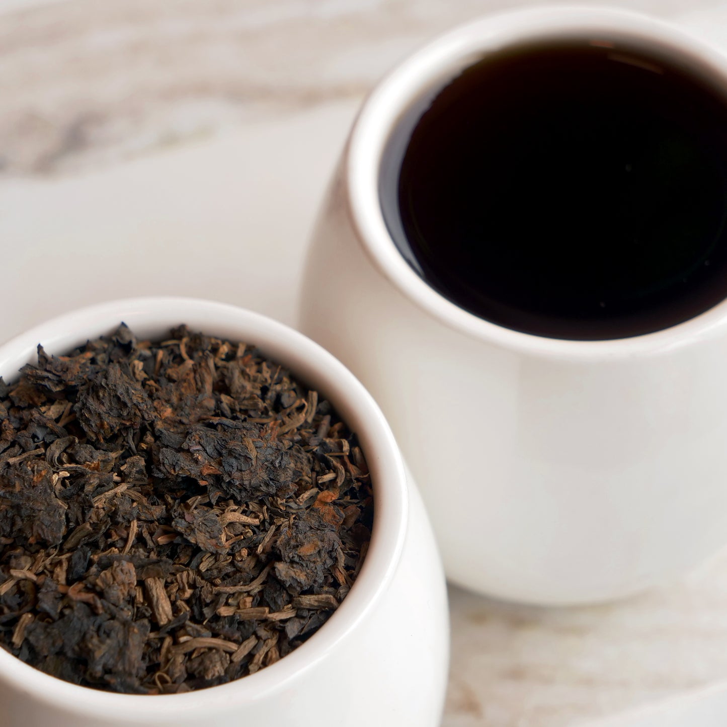 White cup of dark tea next to a bowl of dark tea leaves on a wooden surface