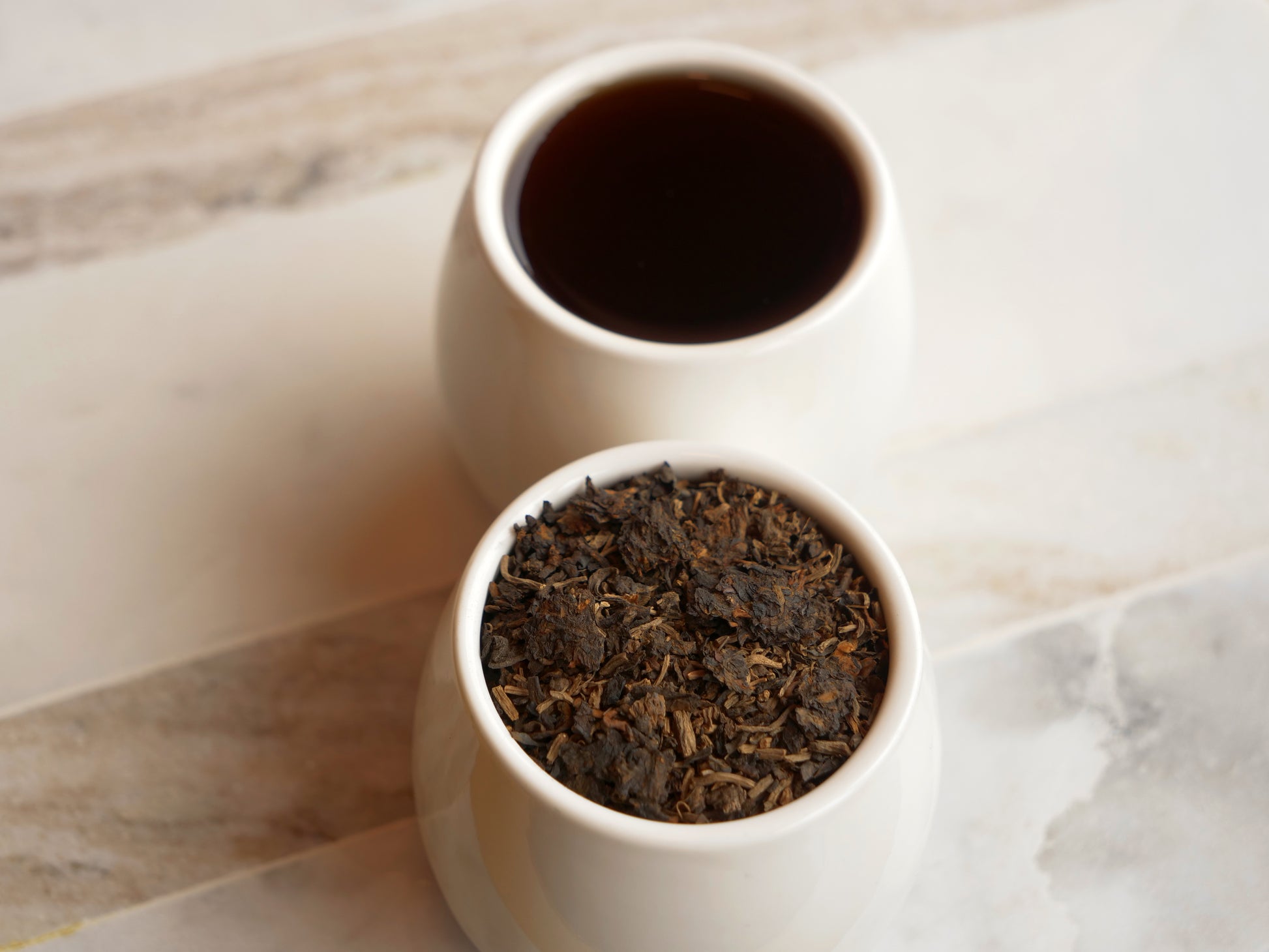 Two white ceramic bowls, one with dark tea and the other with loose tea leaves, on a marble surface.