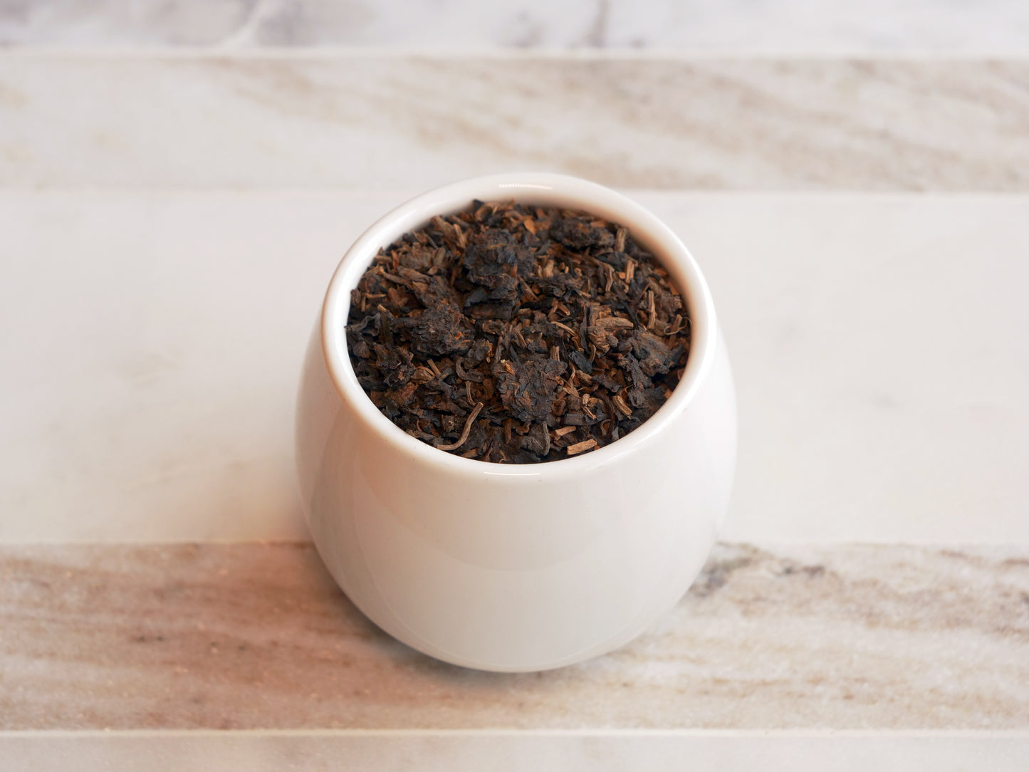 White ceramic bowl filled with dark brown tea leaves on a marble surface