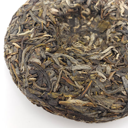 A close-up of a round compressed tea cake made from tightly packed loose-leaf tea, showing dark brown, olive green, and light beige dried leaves, photographed on a white background.