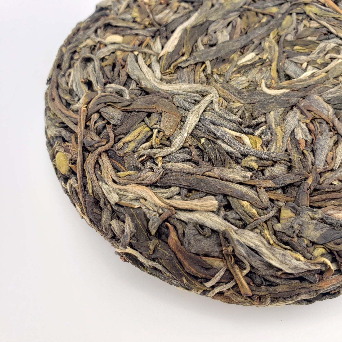 A close-up of a round compressed tea cake made from tightly packed dried tea leaves, showing natural variations in brown, gray, beige, and muted yellow tones, photographed on a plain white background.
