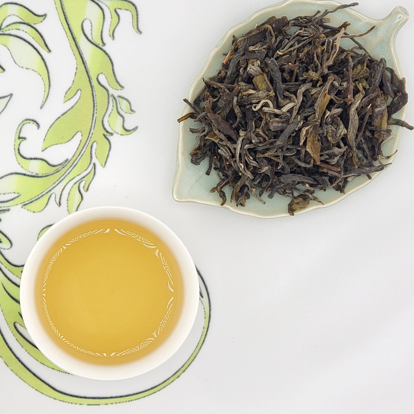 A cup of pale golden tea on a saucer beside a leaf-shaped dish filled with loose dried tea leaves, arranged on a white surface with a green decorative print.