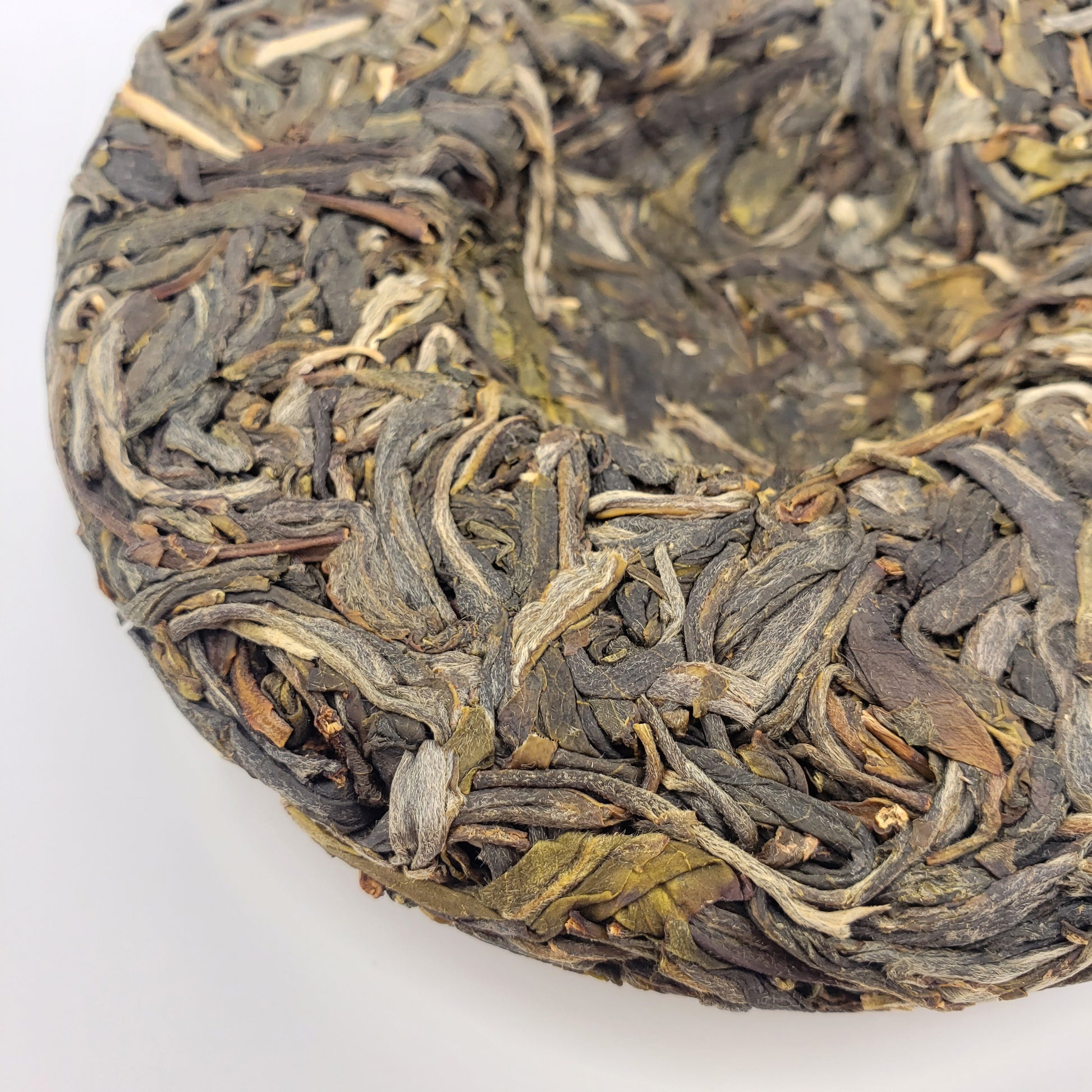 A close-up of a circular compressed tea cake made of tightly packed dried tea leaves, showing varied shades of dark gray, olive green, and brown on a plain white background.