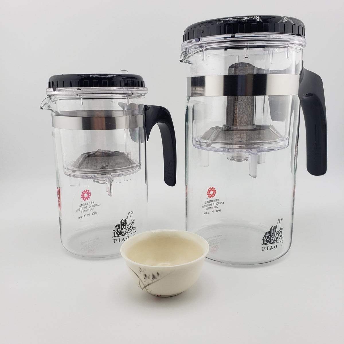 Two clear glass tea infusers with black lids and handles, shown on a white background with a small cream-colored ceramic teacup in front. The brand name 'PIAO I' is visible on the glass.