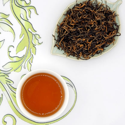 Dried loose leaf tea leaves on a leaf-shaped dish with a cup of brewed auburn tea on a decorative white background.