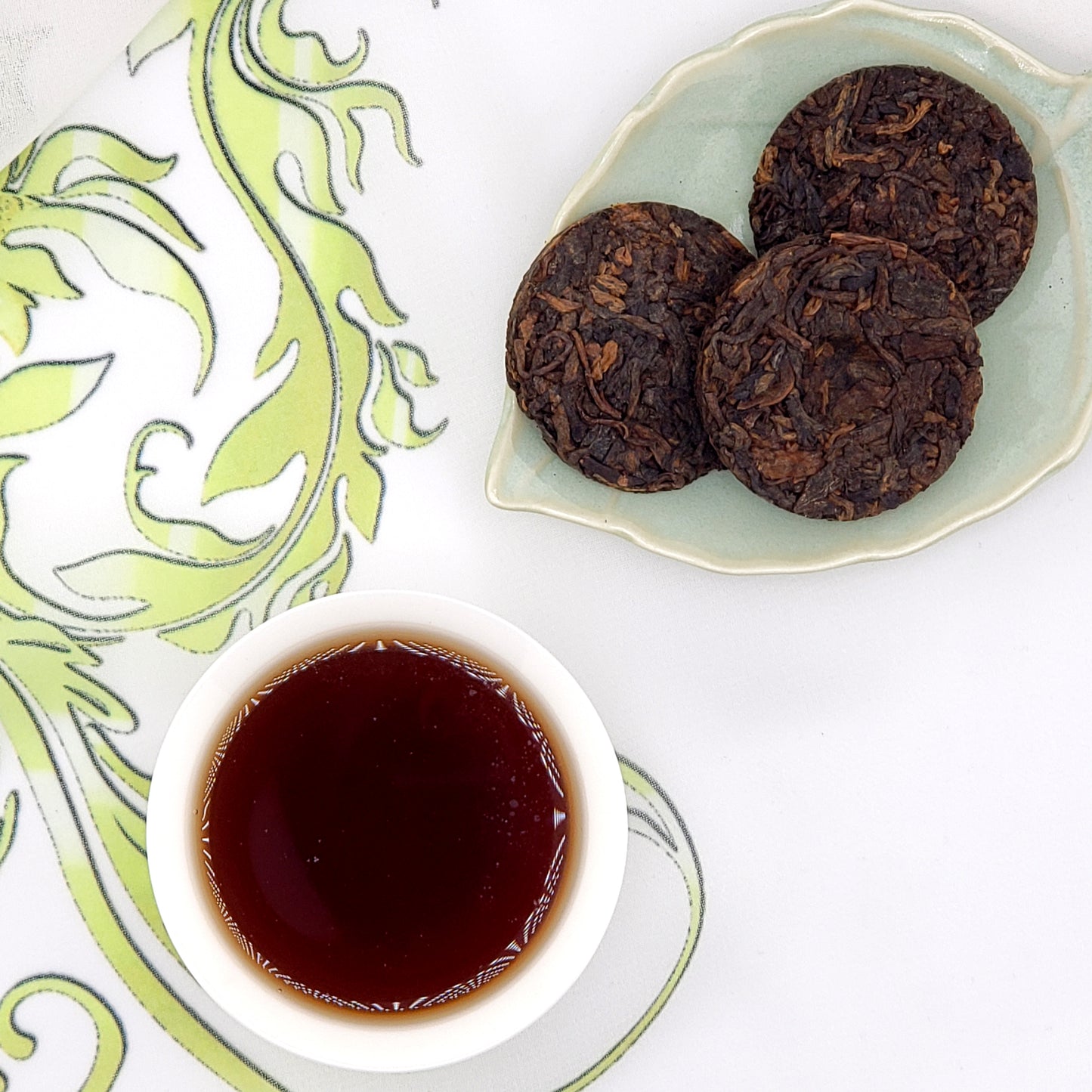 Three dark brown tea coins on a green ceramic plate with a small bowl of brewed tea on a decorative white and green background.