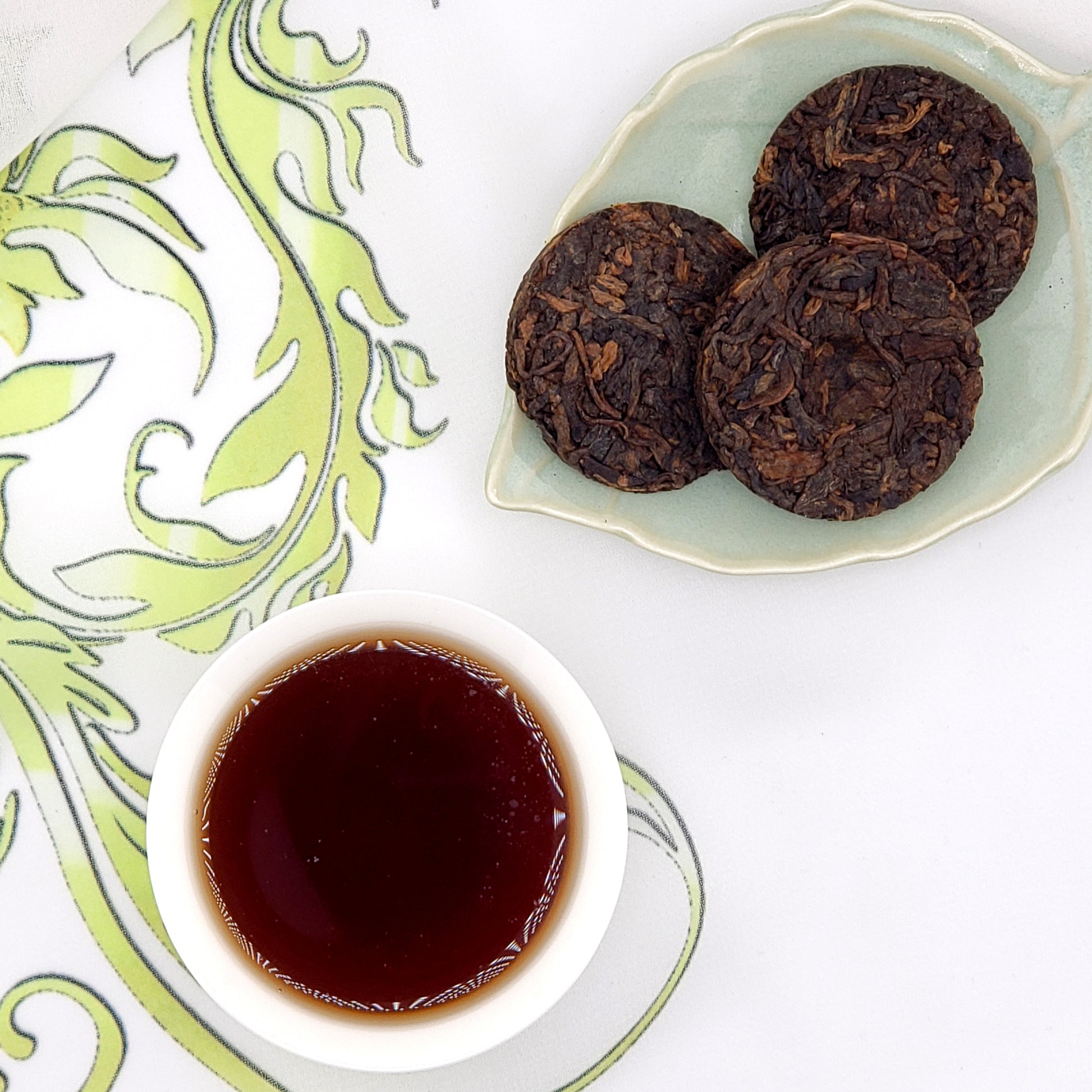 Three dark brown tea coins on a green ceramic plate with a small bowl of brewed tea on a decorative white and green background.