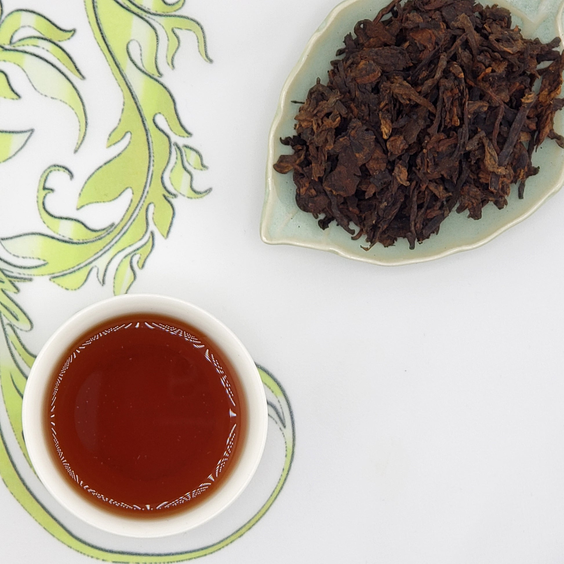 Loose leaf tea leaves on a plate with a cup of rich brown brewed tea on a decorative mat.