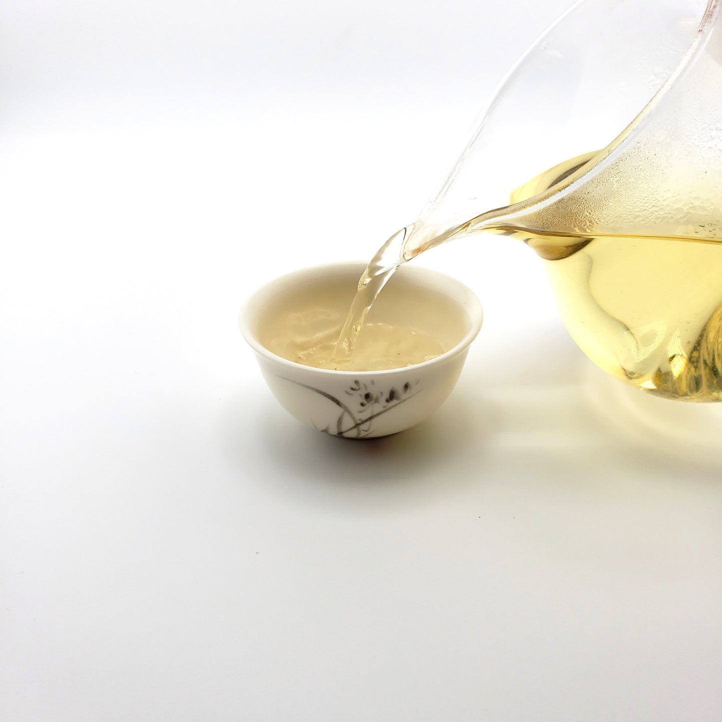 Brewed green tea being poured from a glass pitcher into a small white porcelain cup.