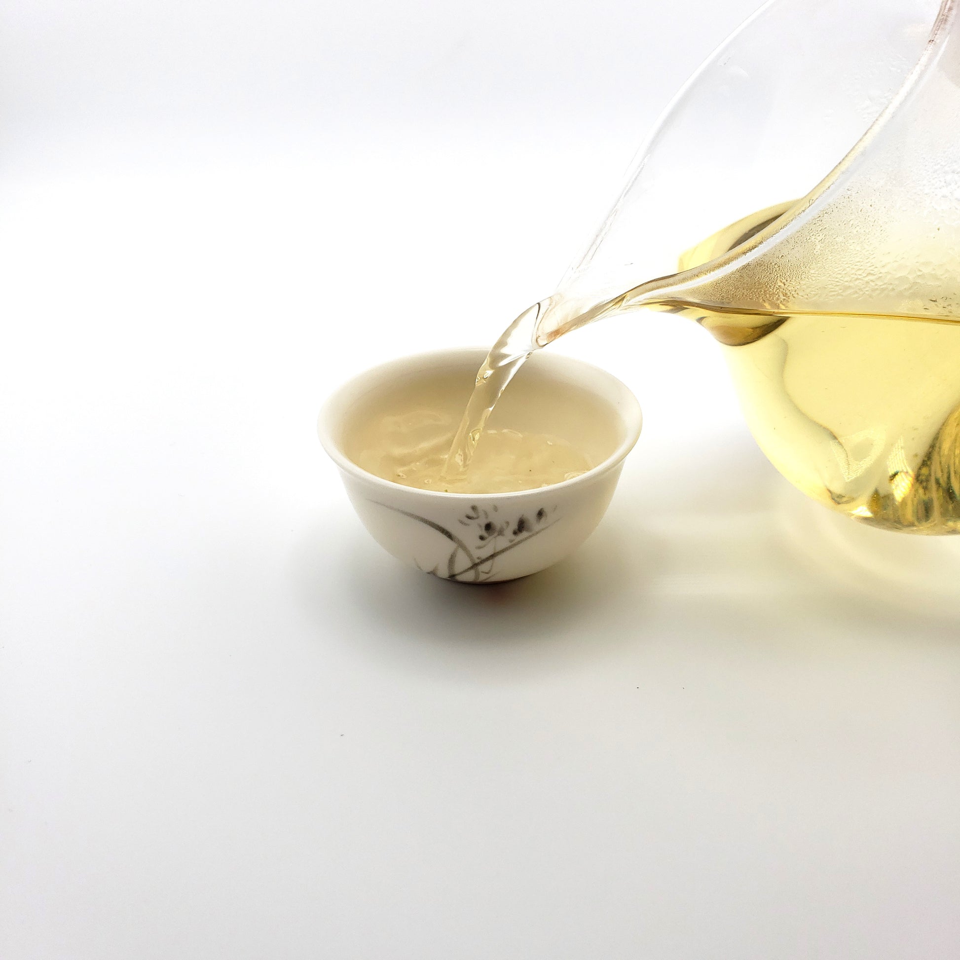 Brewed green tea being poured from a glass pitcher into a small white porcelain cup.