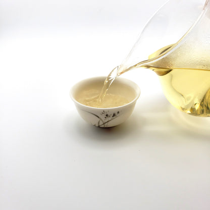 Brewed green tea being poured from a glass pitcher into a small white porcelain cup.