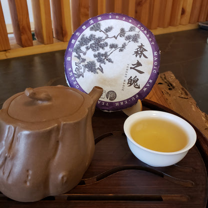 A ceramic tea set arranged on a wooden tray, including a brown clay teapot, a white cup of pale yellow tea, and a round wrapped tea cake with a purple and white label, featuring a tree design and Chinese characters.