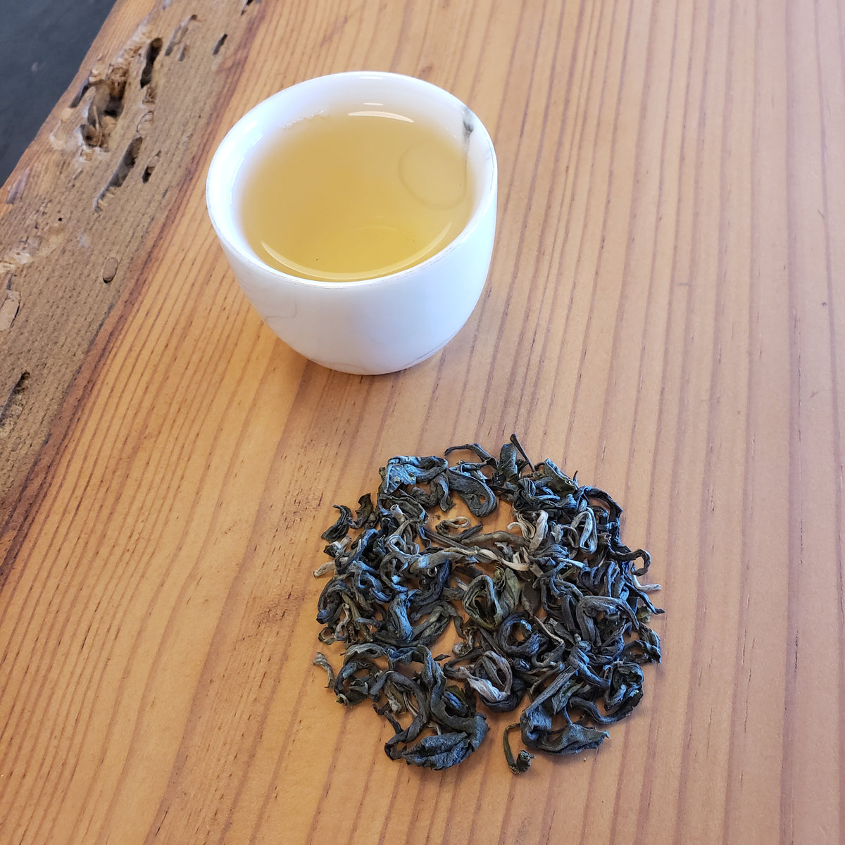 A small white cup of pale yellow tea and a small pile of green loose-leaf tea on a light wood tabletop.