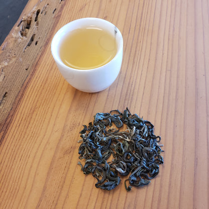 A small white cup of pale yellow tea and a small pile of green loose-leaf tea on a light wood tabletop.